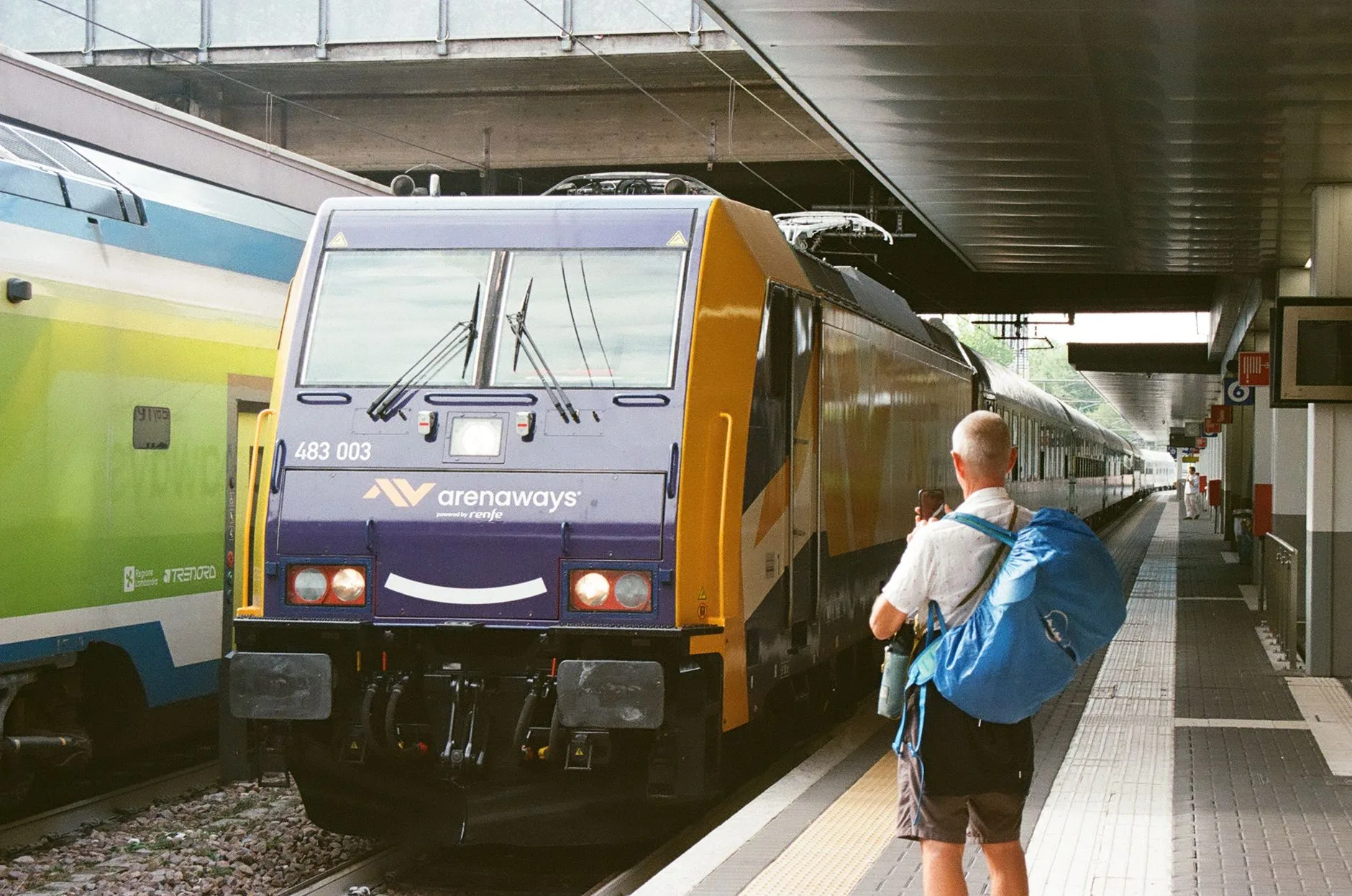 Train speeding past a European platform with mountains and clear sky in the background
